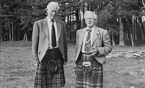 Photo of two men dressed suit jackets, ties and kilts stood on grass before a pine woodland, Colnel John Grant to the left of Sir Patrick Grant
    